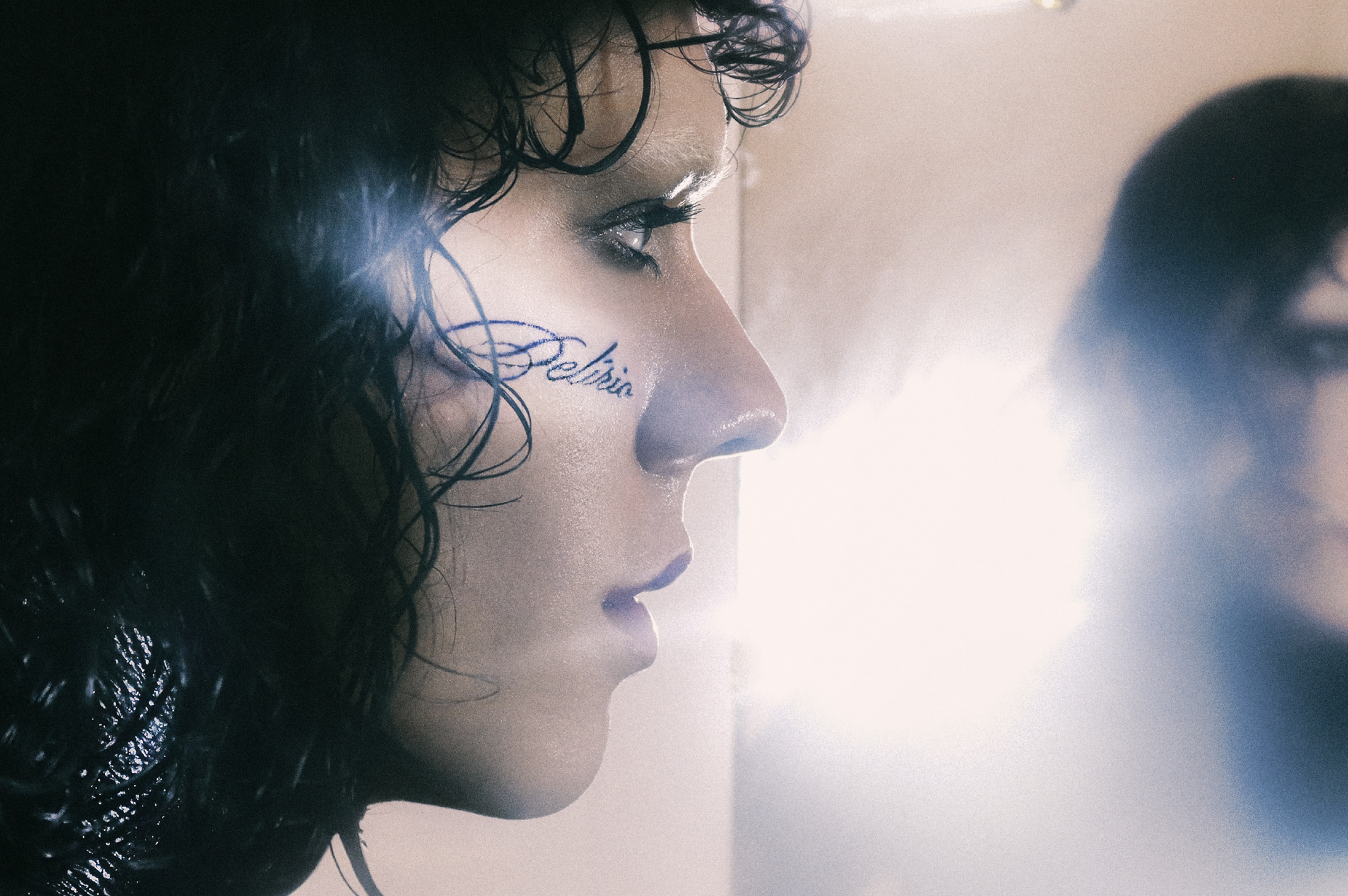 Album cover art showing a closeup of the musician Shi :) looking into a mirror with a light shining, obscuring the reflection slightly. There is a tattoo on the side of their face in cursive that reads "Delirio" - the album title.